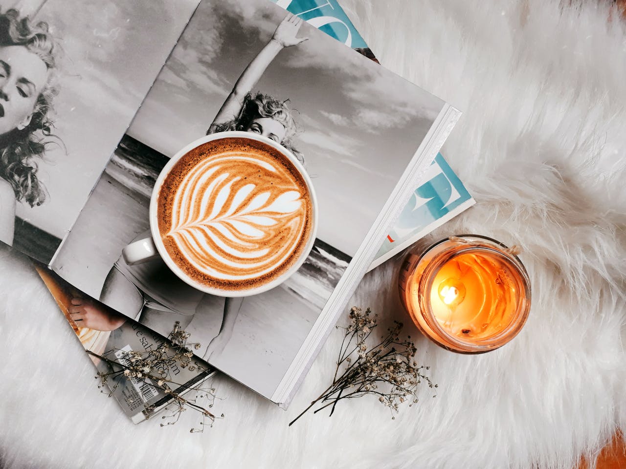 A stylish flat lay featuring a latte with latte art, a magazine, and a candle on a furry surface.
