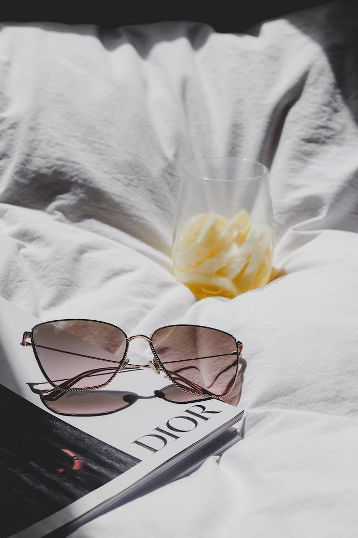 Sunglasses and a refreshing drink on a Dior magazine in soft sunlight, evoking a luxurious and relaxed vibe.